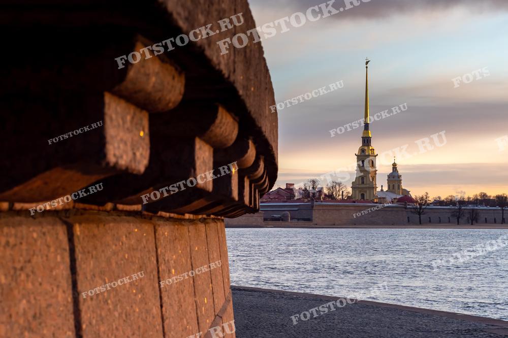 На фотографии представлен вид на Петропавловскую крепость в Санкт-Петербурге, сделанный через реку Неву. Свет заката или рассвета мягко освещает сцену, подчеркивая контуры шпиля с золотым украшением на вершине. Это изображение может быть использовано в исторических книгах, туристических брошюрах, образовательных материалах, а также как часть интерьерного оформления, создающего атмосферу города.